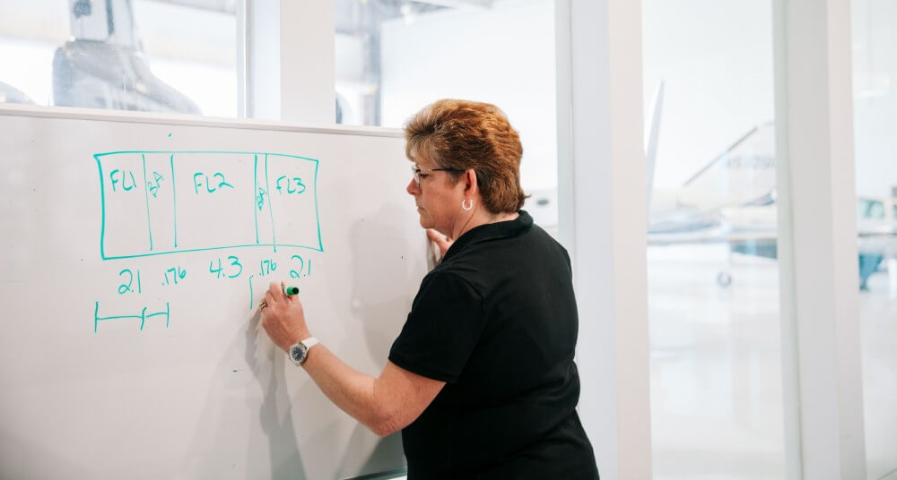 Abingdon Co image displays Dr Gail Rouscher, a woman in her fifties with short brown hair wearing a black short sleeved shirt, is writing on a white board with a green marker. She is teaching fuel systems in a classroom hangar with airplanes in the background