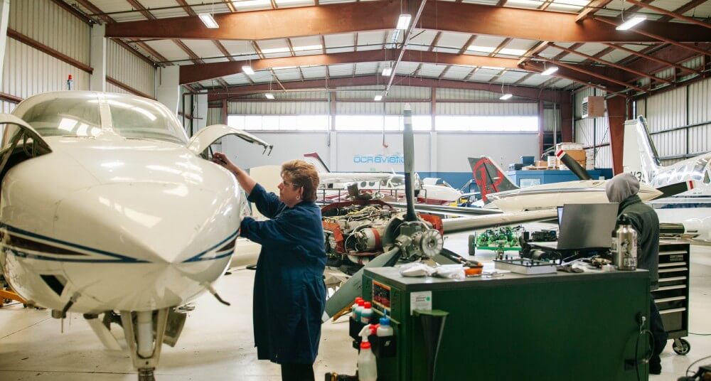 Abingdon Co image displays Dr Gail Rouscher, a woman with short brown hair and wearing blue coverall jacket, working on the fuselage of an airplane in a large aircraft aviation maintenance hangar