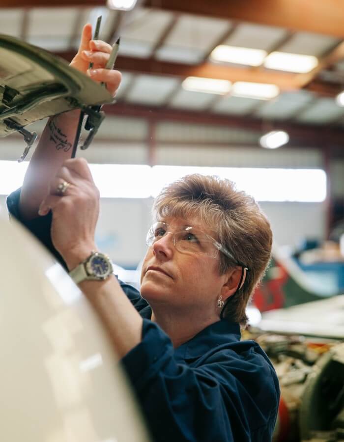 Abingdon Co image displays Dr Gail Rouscher, a woman in her fifties with short brown hair, in a large aircraft hangar inspecting a baggage compartment from the cowl of an airplane. She is wearing safety glasses and is wearing a mechanics coverall. 