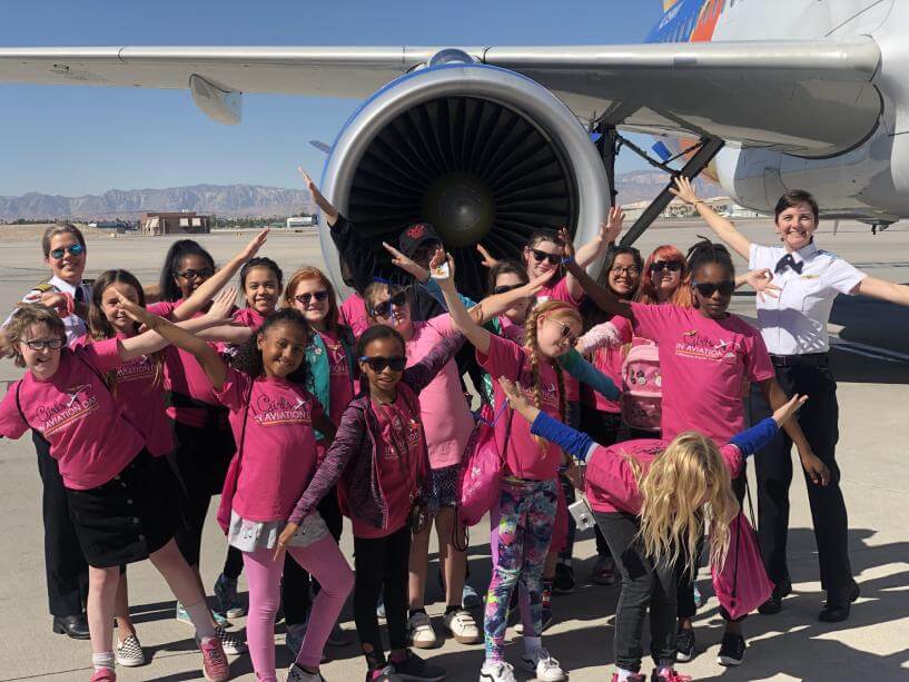 Abingdon Co. Image displays For Girls in Aviation Day, a female Pilot and 18 little girls standing in front of a get engine on an Airbus jet at the airport. All girls have their arms out like airplane wings.