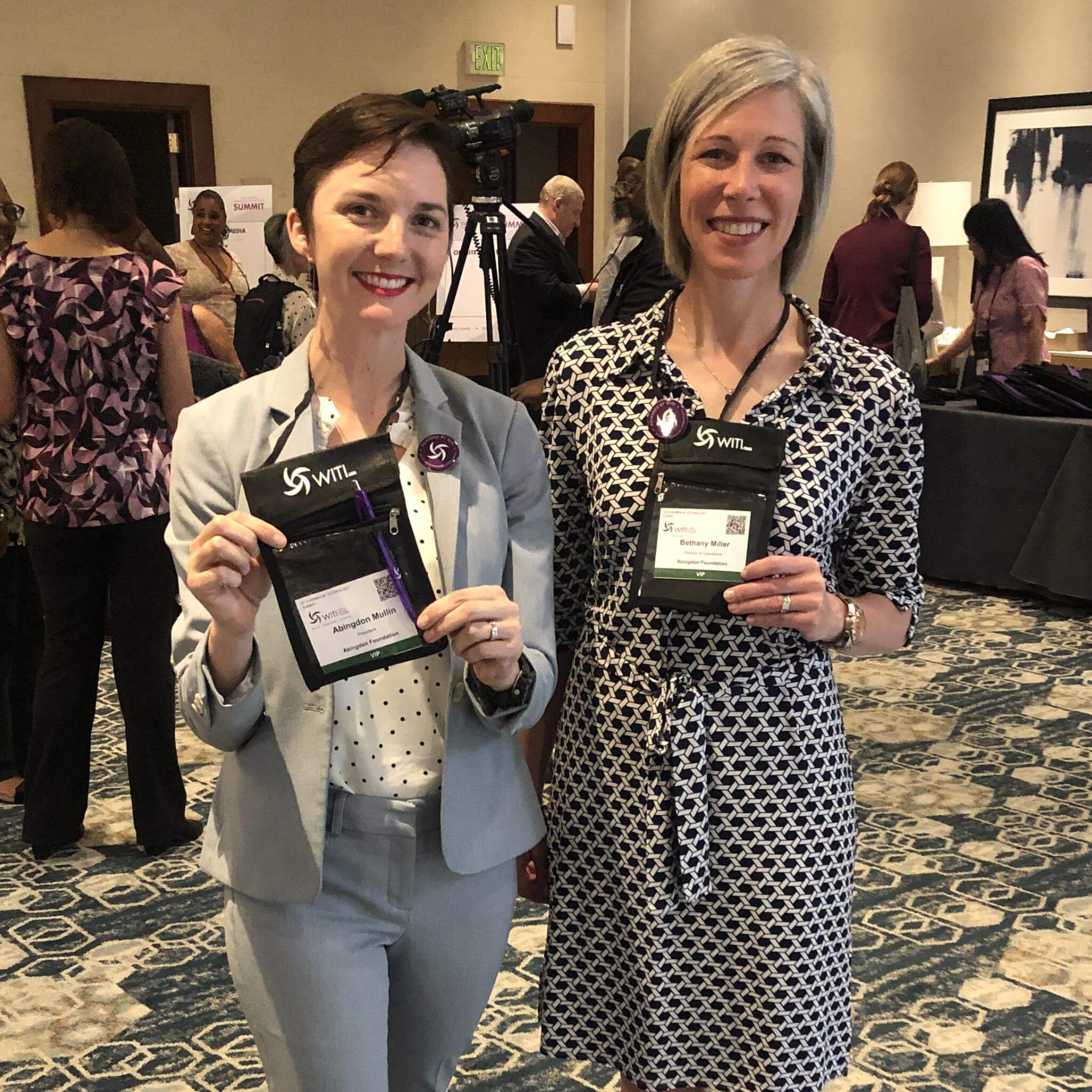 Abingdon Co. Image displays Abingdon Mullin and Bethany Miller representing Abingdon Foundation at Women In Technology International Conference. Both women are standing in a conference lobby holding their name badges to the camera. WITI