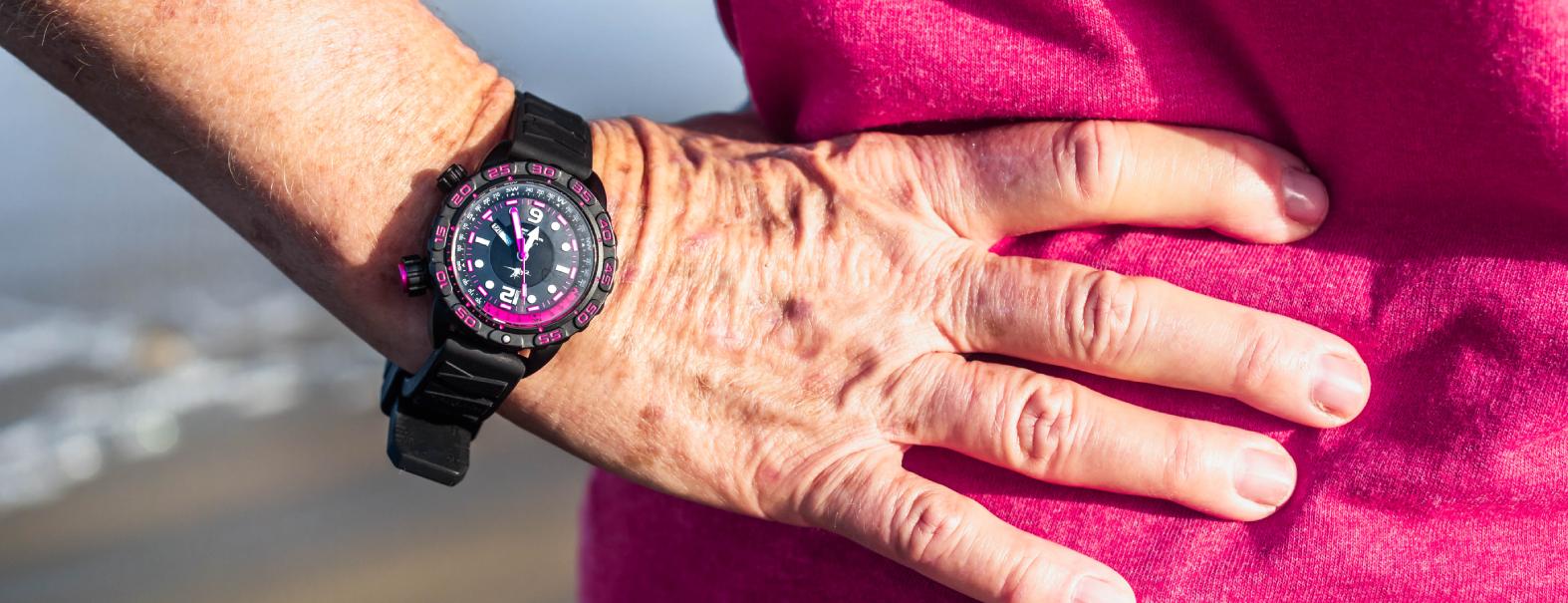 Abingdon Co. A image displaying  Nadia Abyss Compass automatic dive watch with black silicone strap side front view black-pink dial watch on a person wrist with their hand resting on there torso in a closeup pic with a beach back ground