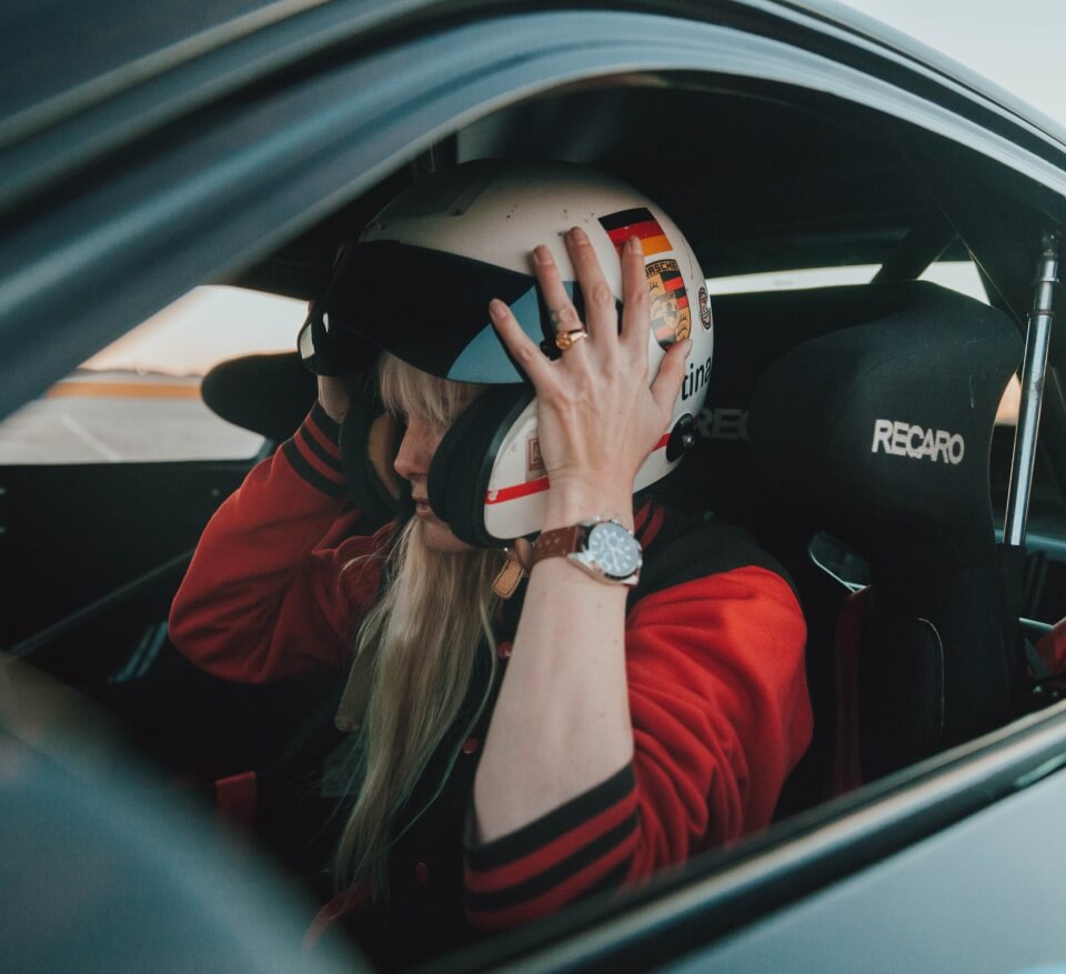 Blonde woman sitting in a race car wearing a red athletic jacket putting on a helmet over her head.