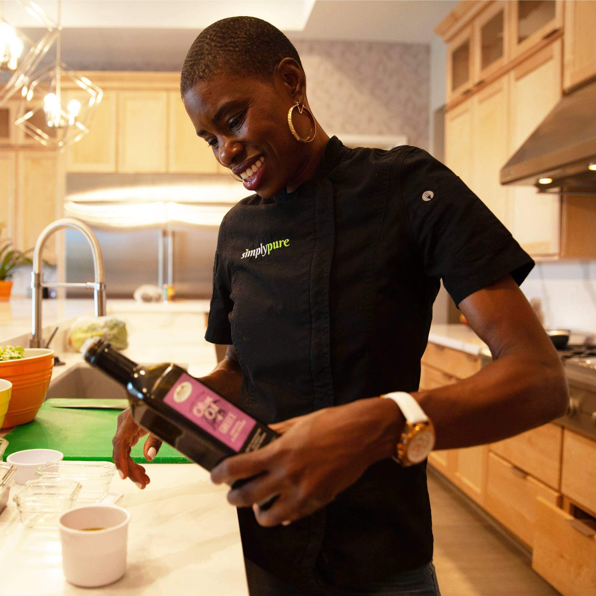 Chef Stacey Dougan, smiling and pouring cooking oil into a small cup in a light wood kitchen wearing an Abingdon watch. 