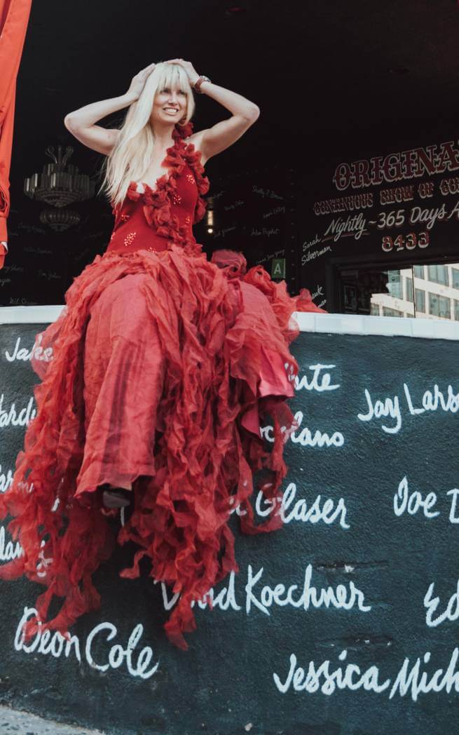 Abingdon Co. image Displays Eugenia Kuzima, model and actress, blonde woman wearing a long red dress sitting on a ledge with her arms on her head. Daytime and she is smiling and looking off in the distance