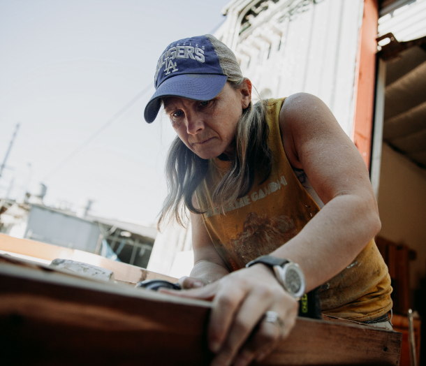 Abingdon Co. A image displaying Carpenter Marjorie works outside in her workshop with a piece of wood wearing a baseball cap and a black leather band watch.
