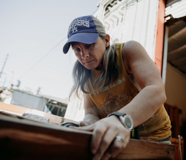 Abingdon Co. A image displaying  Carpenter Marjorie works outside in her workshop with a piece of wood wearing a baseball cap and a black leather band watch.