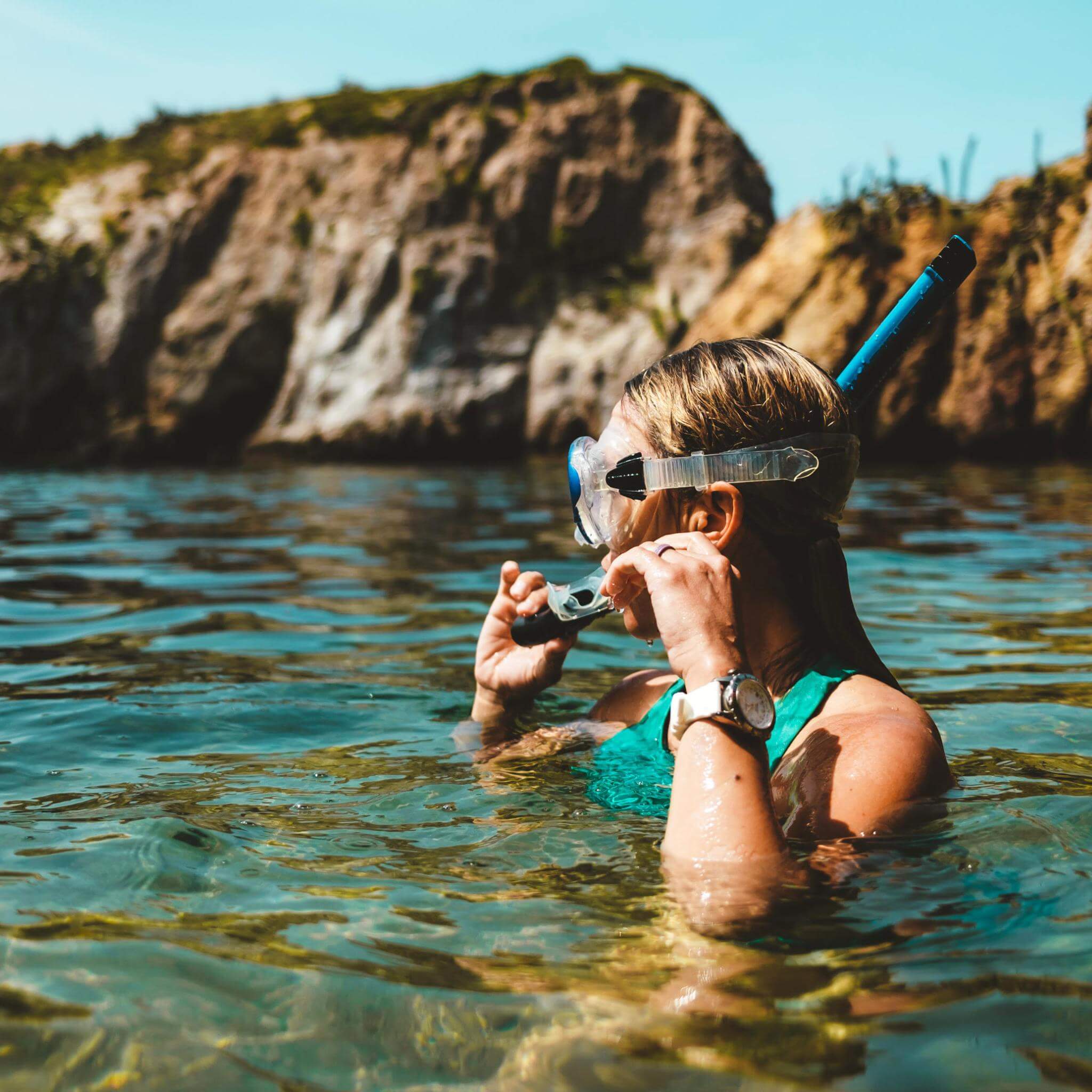 Woman wearing a snorkel mask in the ocean holding the mouthpiece above the water with rocks in the background