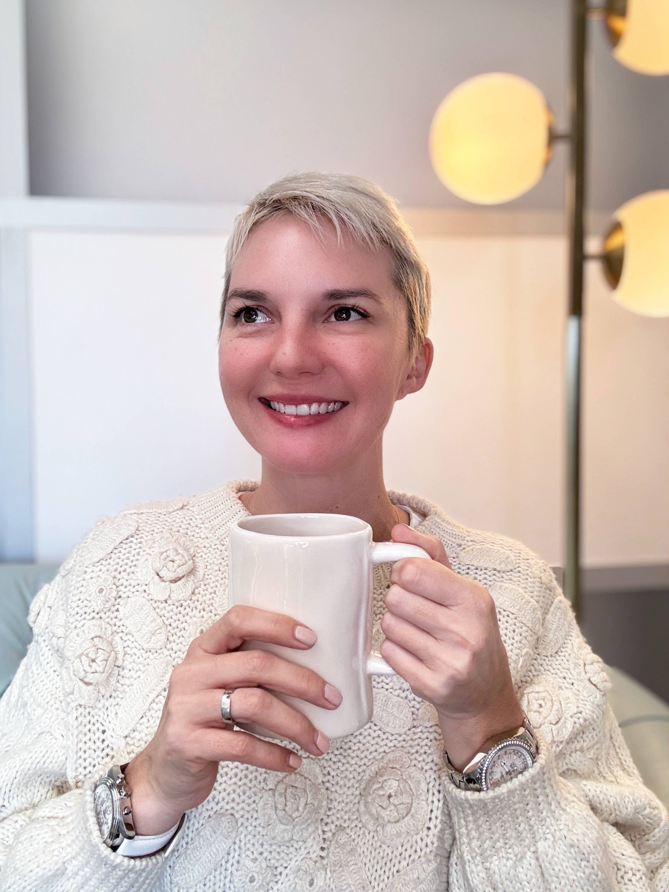 Abingdon Mullin, a middle aged woman with short blonde hair wearing a white sweater is holding a coffee mug with both hands. She has two watches on her wrists and is looking up and to the left. A lamp and wall are behind her