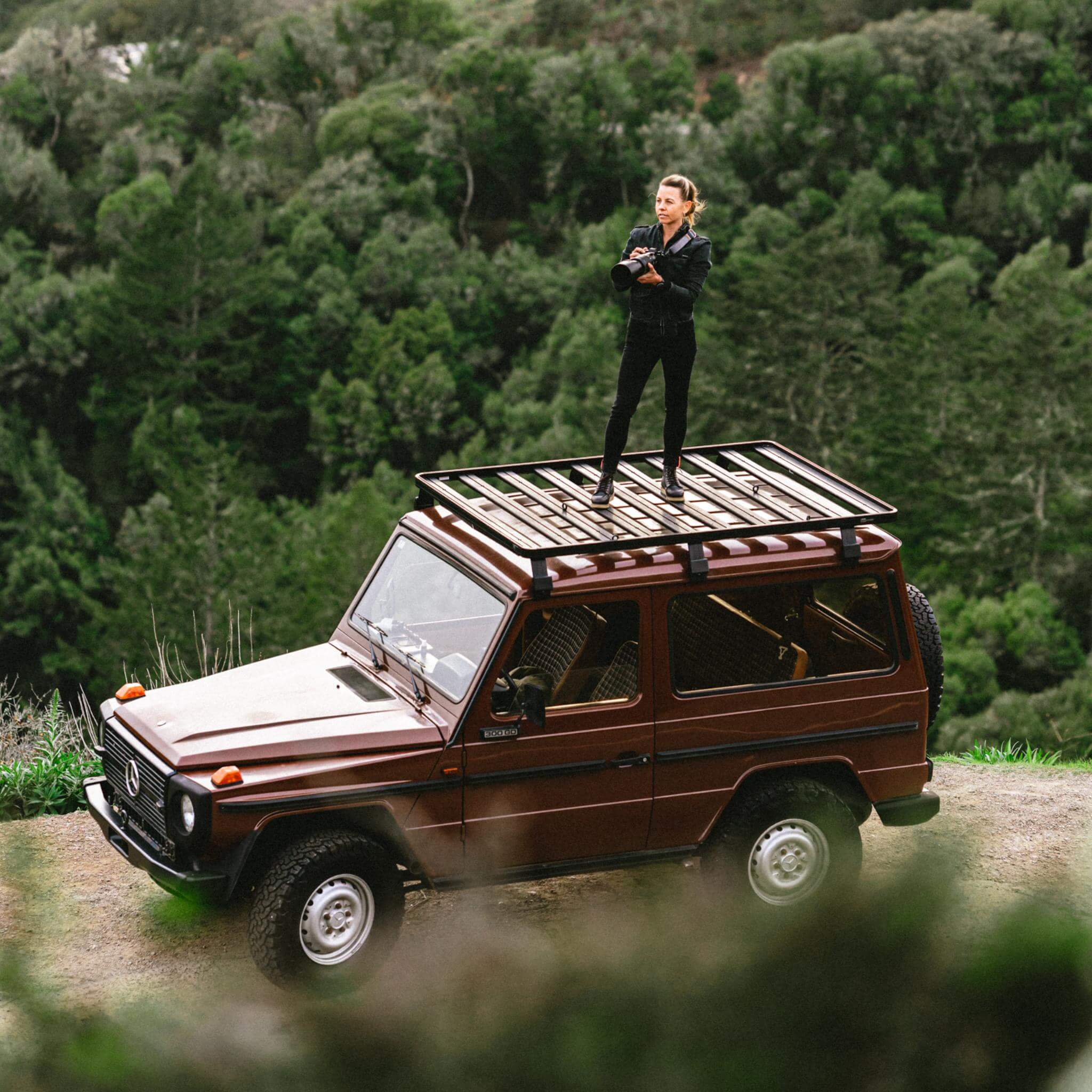 Abingdon Co. Image Displays Woman standing on roof of red mercedes outside in nature holding a camera. 