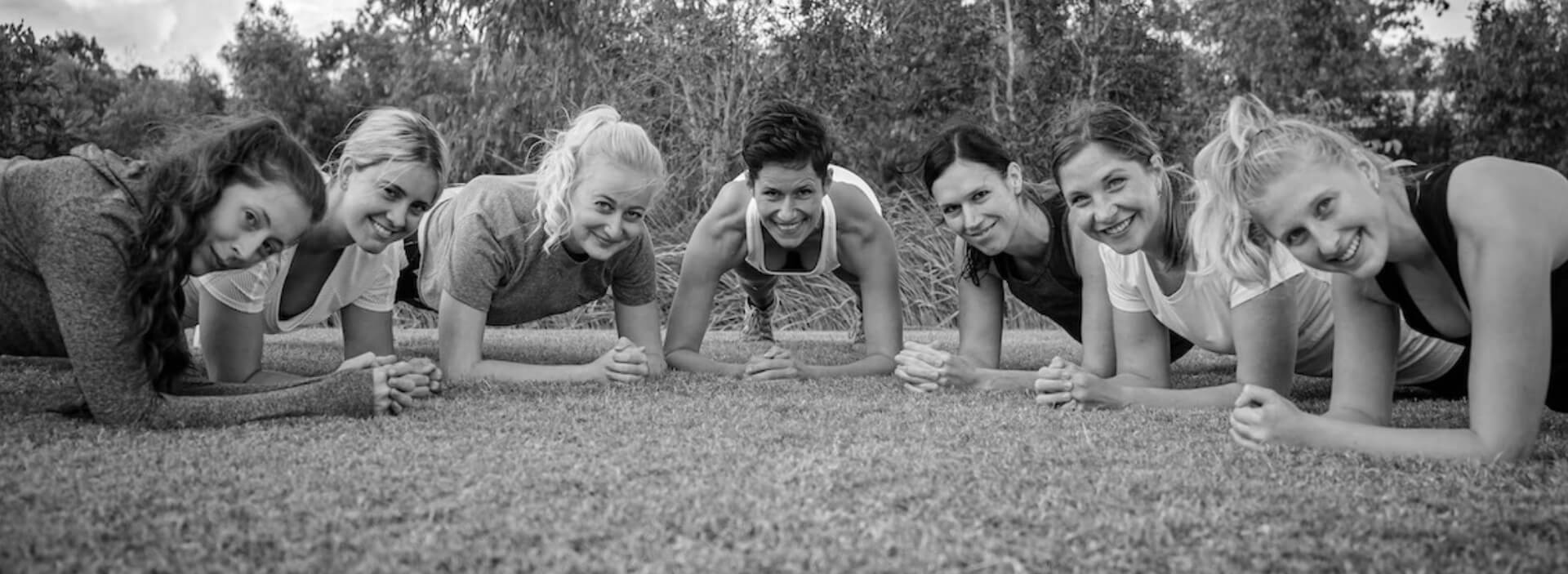 Abingdon Co. image displays a diverse group of individual woman all in a pushup position besides another in a line Smiling and looking straight at the camera in a forest setting while on the grass this picture is in black and white.