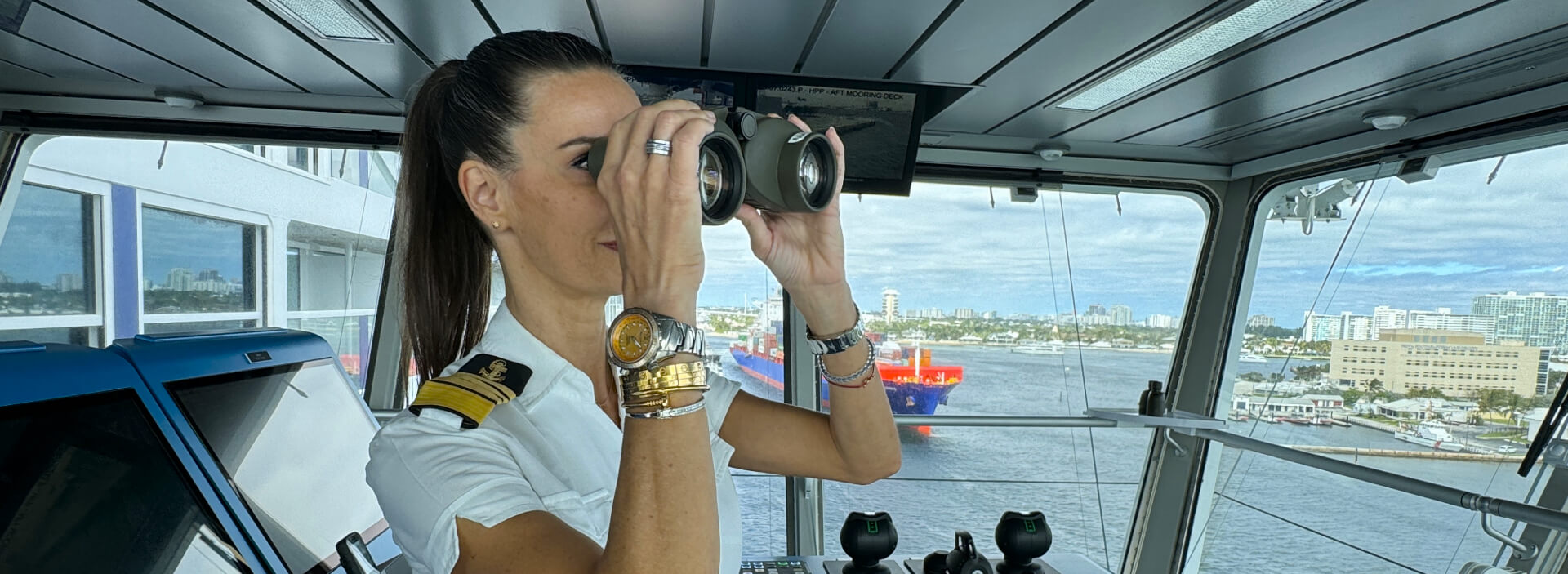 Captain Kate McCue, looks through a pair of binoculars while on the bridge of Celebrity Beyond, the mega cruise ship she leads. Captain Kate has on her captain uniform, with her hair pulled back in a ponytail and her Abingdon watch on her wrist.