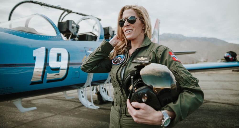 Abingdon Co image of Stephanie Goetz standing in front of her L-39 Albatross jet smiling and holding her helmet. She is a blonde woman wearing aviator sunglasses and a military green flight suit with patches on it. 