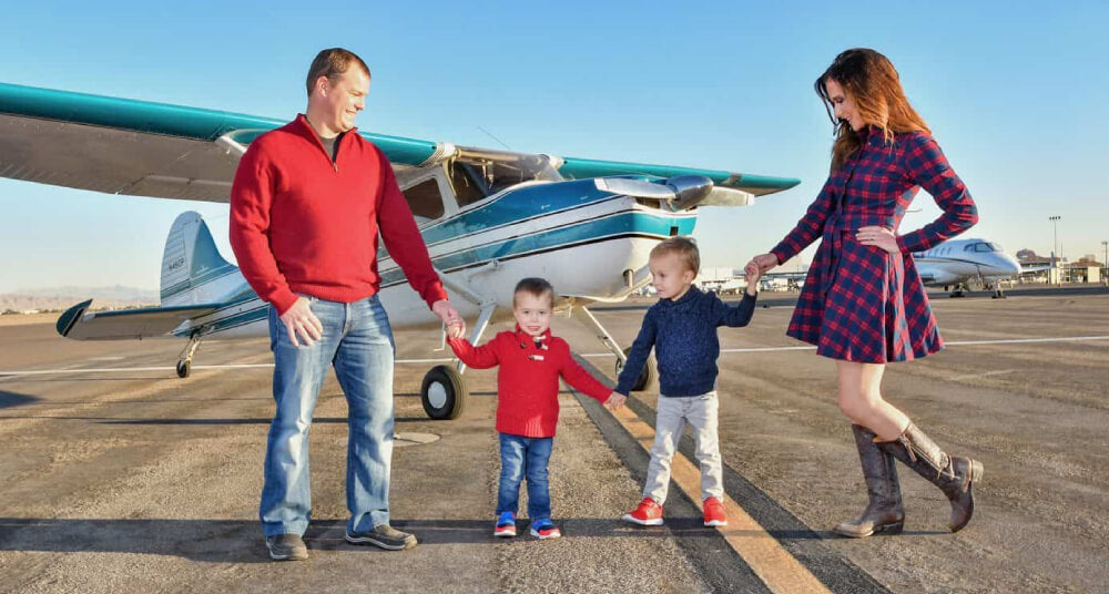 A family of four, husband, two young boys, and wife, all stand holding hands on the pavement at the airport in front of a blue and white vintage Cessna airplane. Both parents are looking down at their children. It is sunny outside and the father and boys are wearing jeans and long plain sweaters while the mother is wearing a blue and red plaid dress with long brown boots. It is a picture of a happy family.