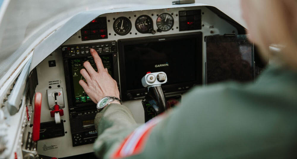 Abingdon Co image of the panel of a L-39 Albatross jet where a woman's hand is manipulating the instruments. The woman is wearing a green flight suit and a green watch and is pressing buttons on the instrument panel. 