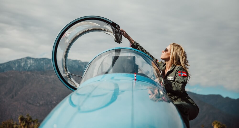 Abingdon Co image of Stephanie Goetz standing on the steps of her L-39 Albatross jet reaching above her to pull the canopy down. She is a blonde woman wearing aviator sunglasses and a military green flight suit with patches on it. 