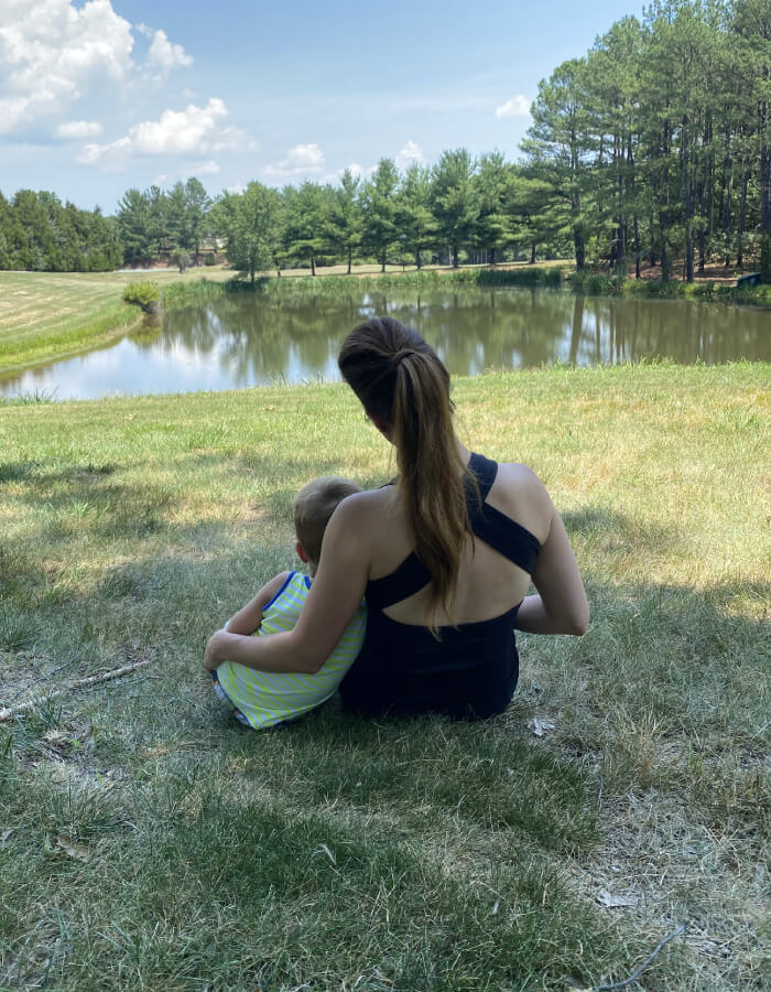 A woman and child are sitting on the grass by a lake. The woman is wearing a black top and has her left arm around the boy who is leaning into his mother. Both mother and child are facing away from the camera.