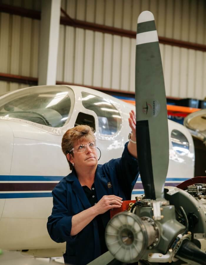 Abingdon Co image displays Dr Gail Rouscher, a woman in her fifties with short brown hair, in an aircraft hangar examining a propeller on a twin engine airplane. She is wearing safety glasses and has on a dark blue coverall. 