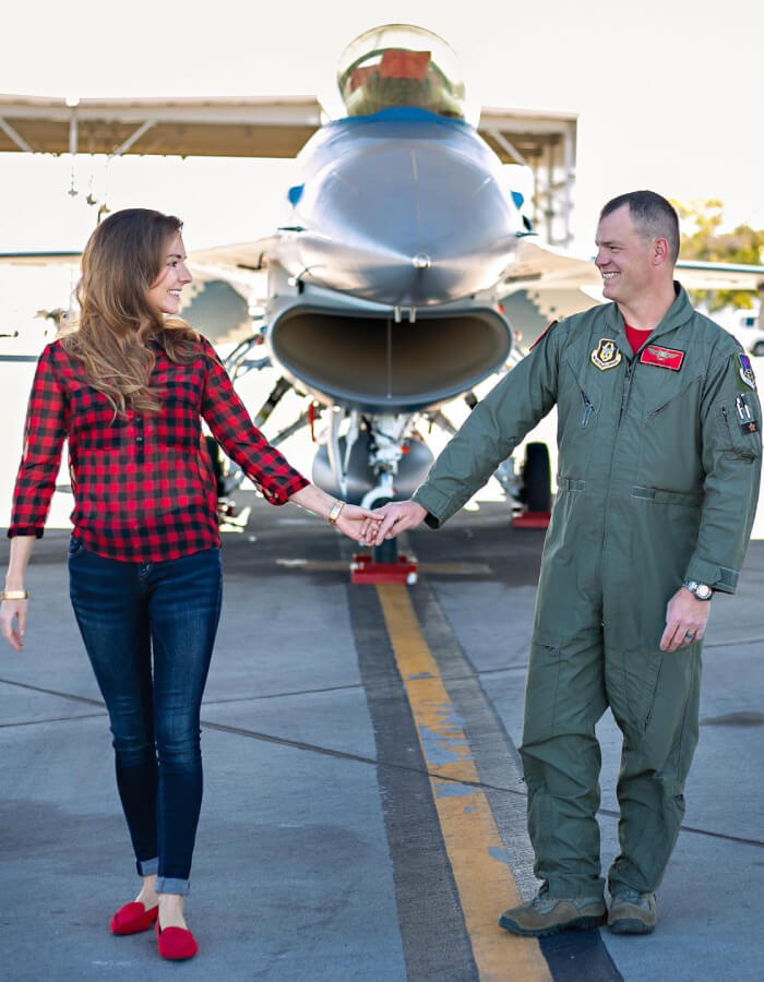 Dianna and Mark Klein are standing and holding hands in front of the nose of a parked jet and looking at each other smiling.