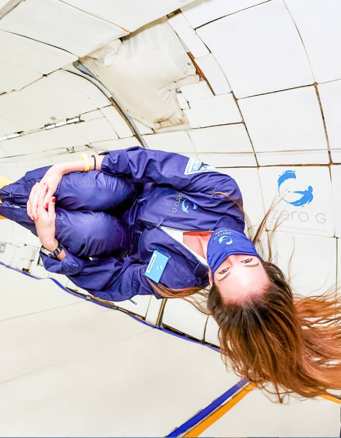 A woman is weightless in the fuselage of an airplane, upside down holding her knees up to her chest, suspended in air. She is wearing a flight suit and a face mask, and her brown hair is flowing all around her.