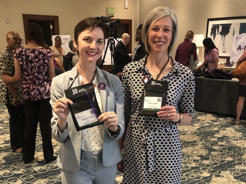 Abingdon Co. Image displays Abingdon Mullin and Bethany Miller representing Abingdon Foundation at Women In Technology International Conference. Both women are standing in a conference lobby holding their name badges to the camera. WITI
