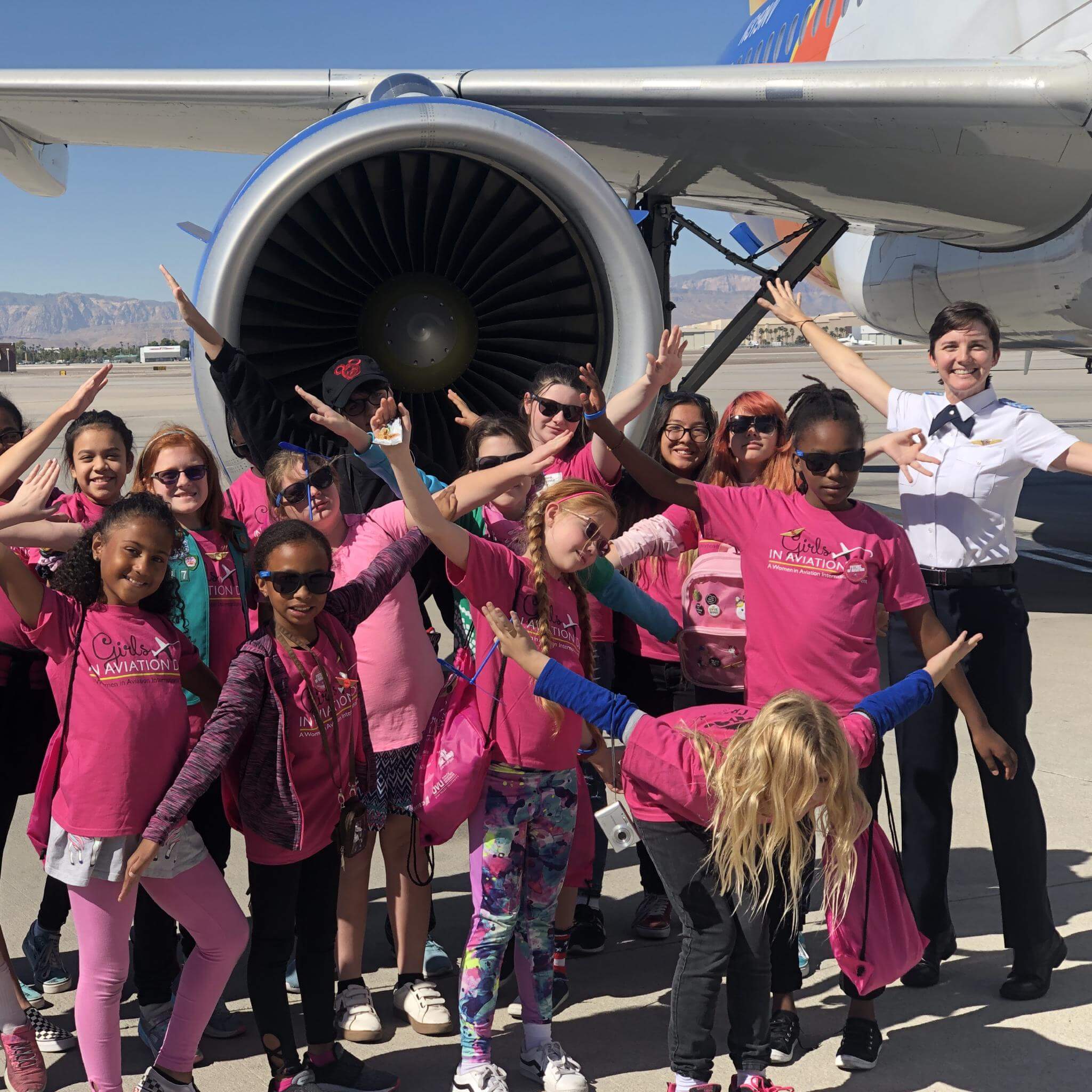 Abingdon Co. Image displays For Girls in Aviation Day, a female Pilot and 18 little girls standing in front of a get engine on an Airbus jet at the airport. All girls have their arms out like airplane wings.