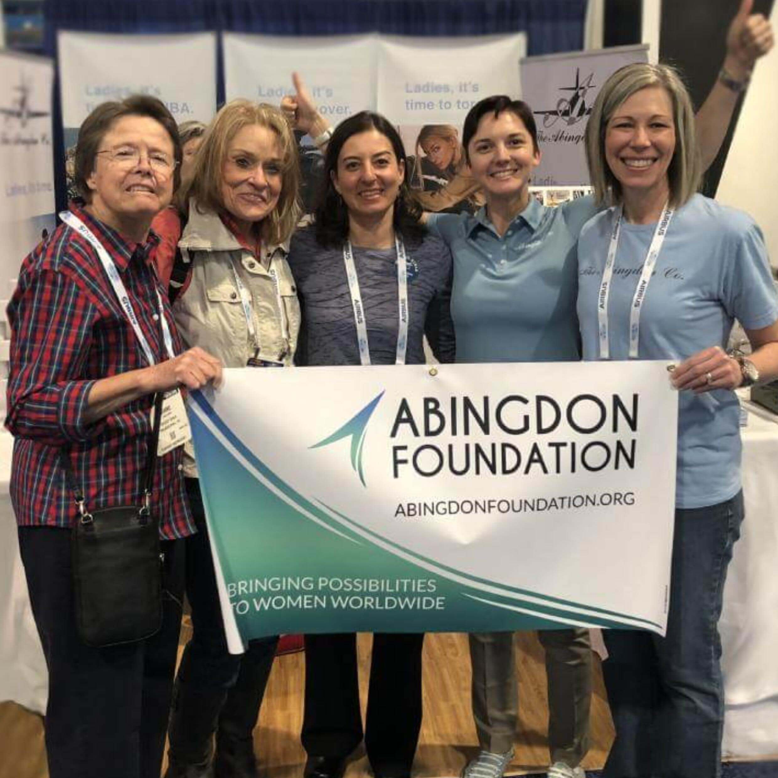 Five women standing at a tradeshow booth holding a banner. The woman in the middle of the five is a scholarship winner who was awarded a fully paid trip to the event. The four other women are mentors and advisors. 
