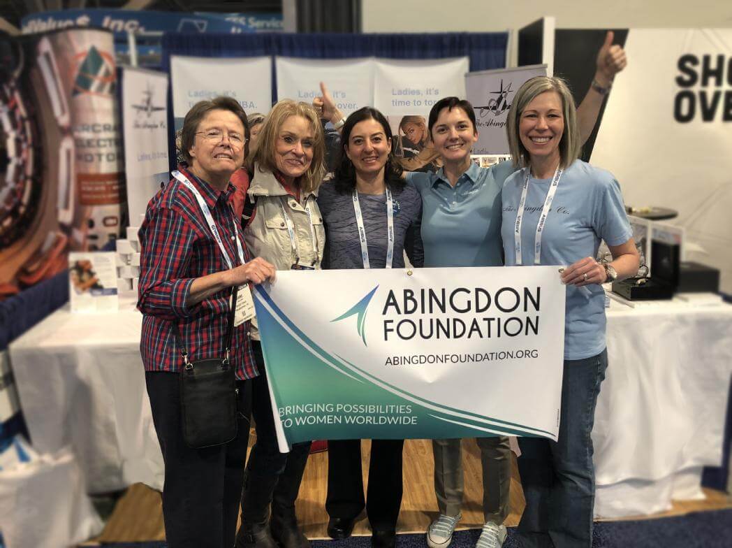 Abingdon Co. Image displays Five women standing at a tradeshow booth holding a banner. The woman in the middle of the five is a scholarship winner who was awarded a fully paid trip to the event. The four other women are mentors and advisors. 