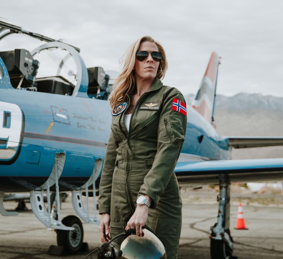 Abingdon Co image of Stephanie Goetz standing in front of her L-39 Albatross jet holding her helmet. She is a blonde woman wearing aviator sunglasses and a military green flight suit with patches on it. 