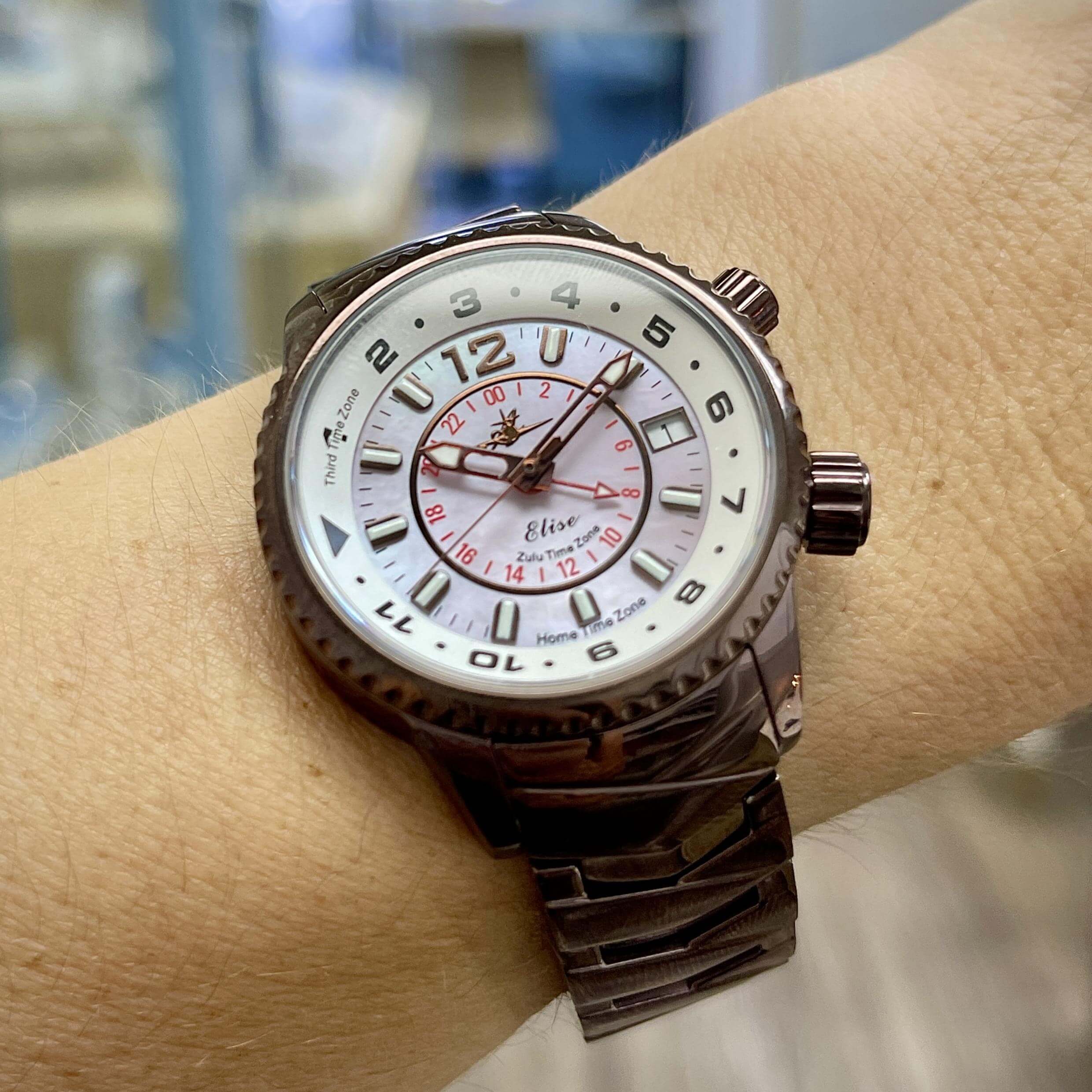 Abingdon Co. Image displays Elise brown travel watch with pink dial on a wrist indoor shot with furniture in the background. Elise travel watch has three time zones displayed on a single circular dial. Mother of pearl face and GMT displayed women's watch