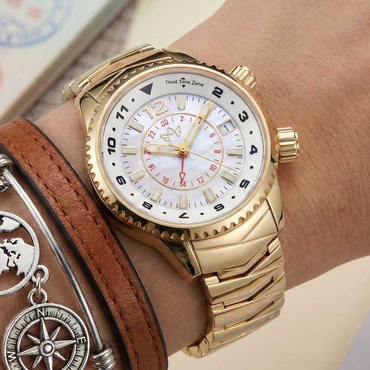Abingdon Elise gold wristwatch with detailed face on a wrist, stacked with a brown leather strap and silver charms.