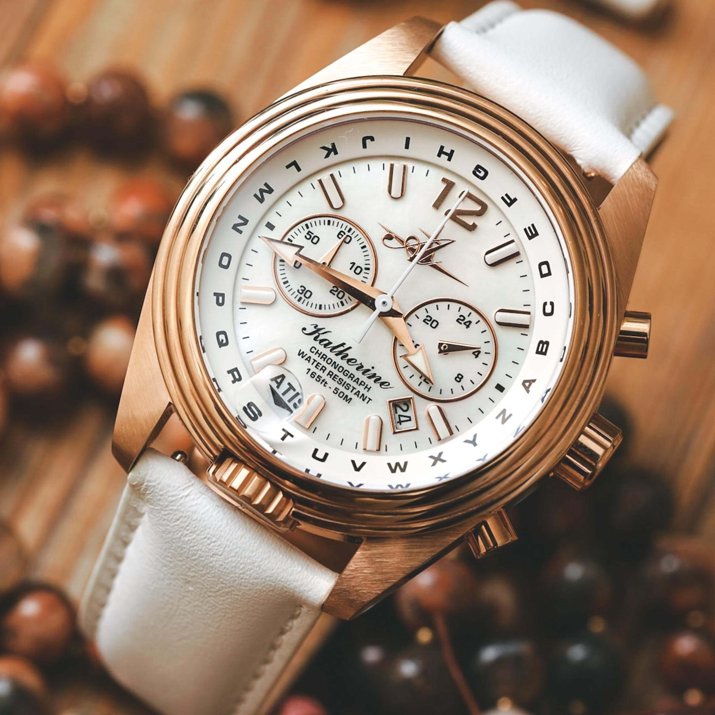 Abingdon Co. Image displays Katherine watch with rose gold case and white leather band in front of a background with beads and wood. White pearl dial with stopwatch, date display, and military time. Pilot aviation watch for women by Abingdon Company