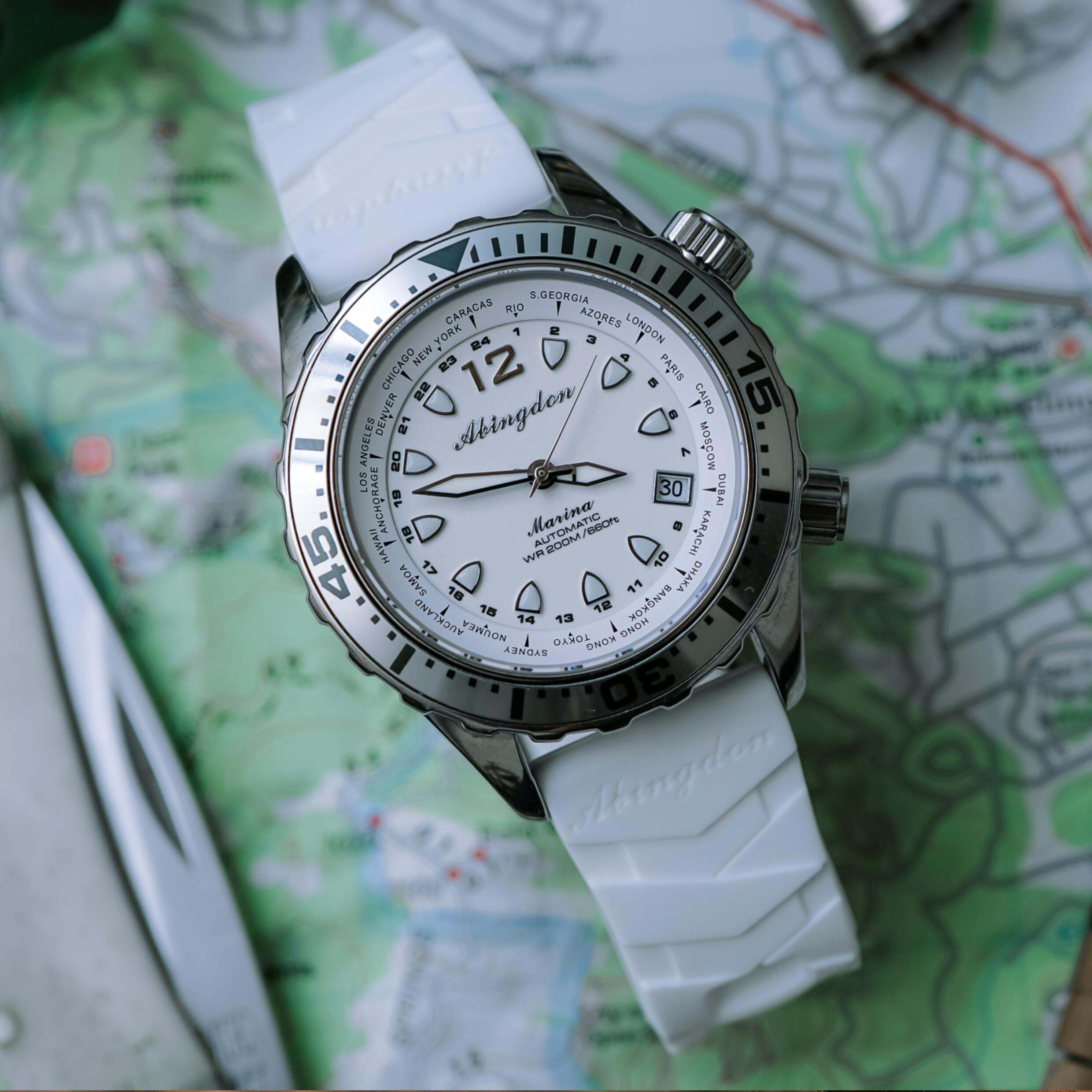 Abingdon Co. Image displays Marina divers watch with white silicone strap laying flat on a topographical chart with a pocketknife next to it. White dial, white hands that glow in the dark, the Marina automatic watch is designed for SCUBA divers and is 660 feet water resistant