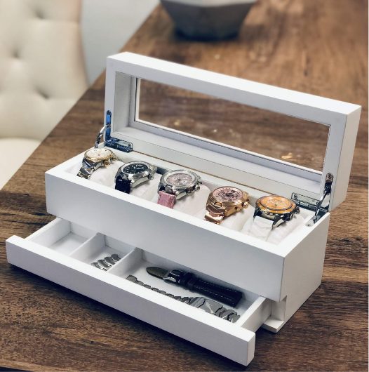 Abingdon Co. image displaying white watch collection box open and sitting on a wooden desk. Five watches and two bands are displayed inside the collector box. Caywood Collectors box has a glass top and white felt pillows.