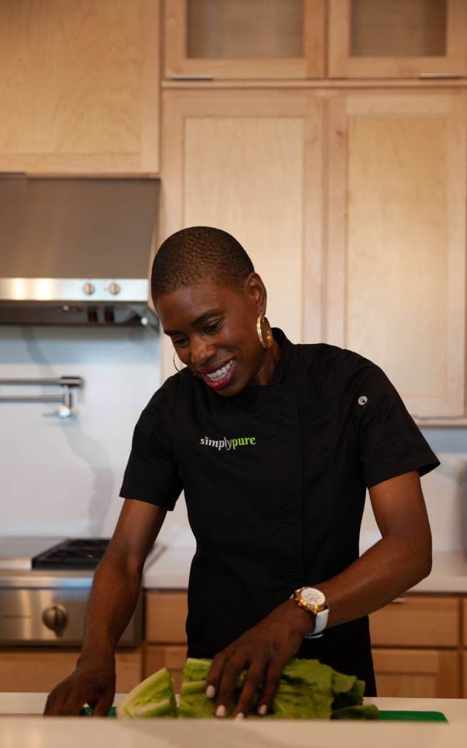 Chef Stacey Dougan, smiling and cutting lettuce in a light wood kitchen wearing an Abingdon watch. 