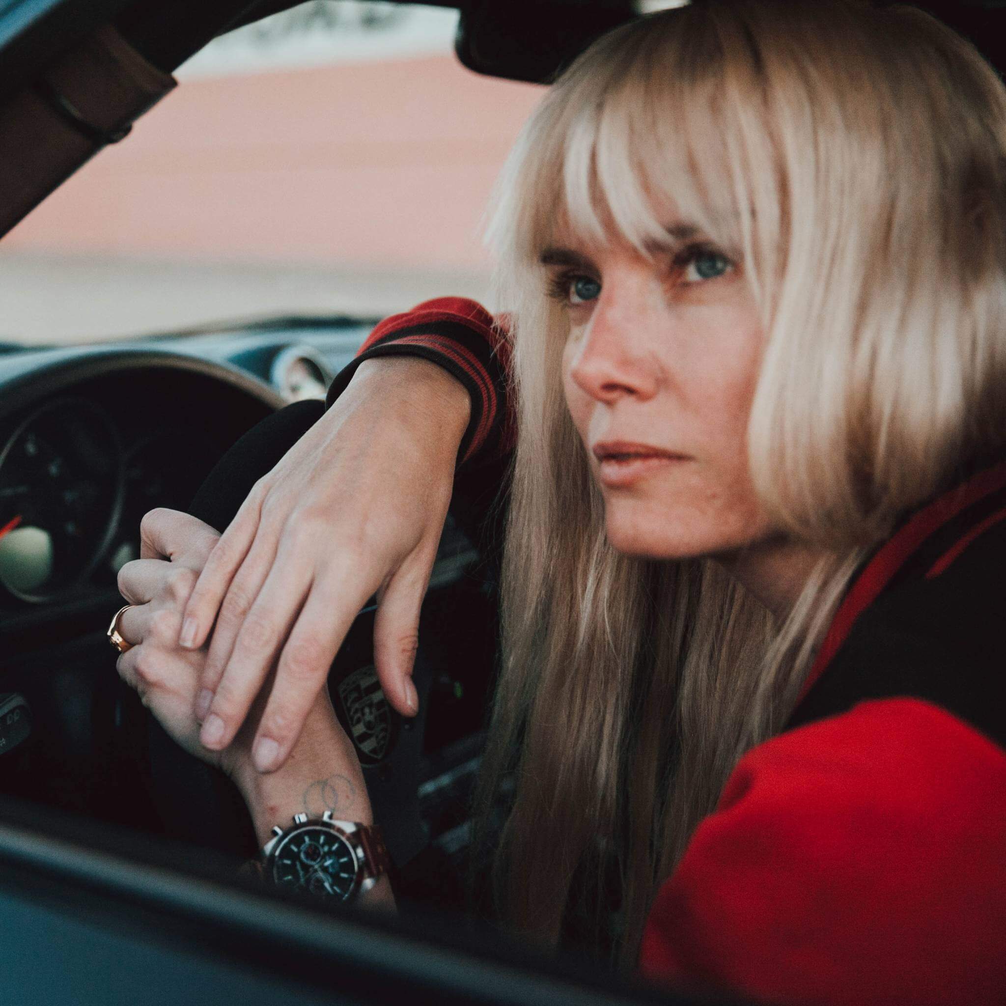 Abingdon Co. Image Displays Model actress, Eugenia Kuzmina sitting in a race car with her hands on the wheel looking out the side wearing a red and black jacket