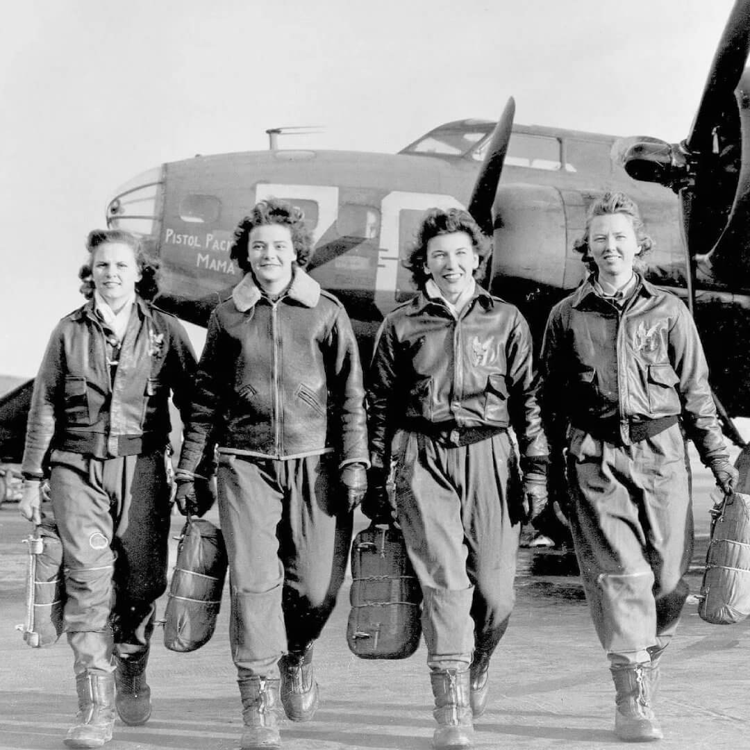 Black and white historical image of four women walking shoulder to shoulder towards the camera with a vintage airplane behind them. Each woman is smiling, wearing a leather bomber jacket, pants, and carrying a bag in their hands, possibly a parachute.